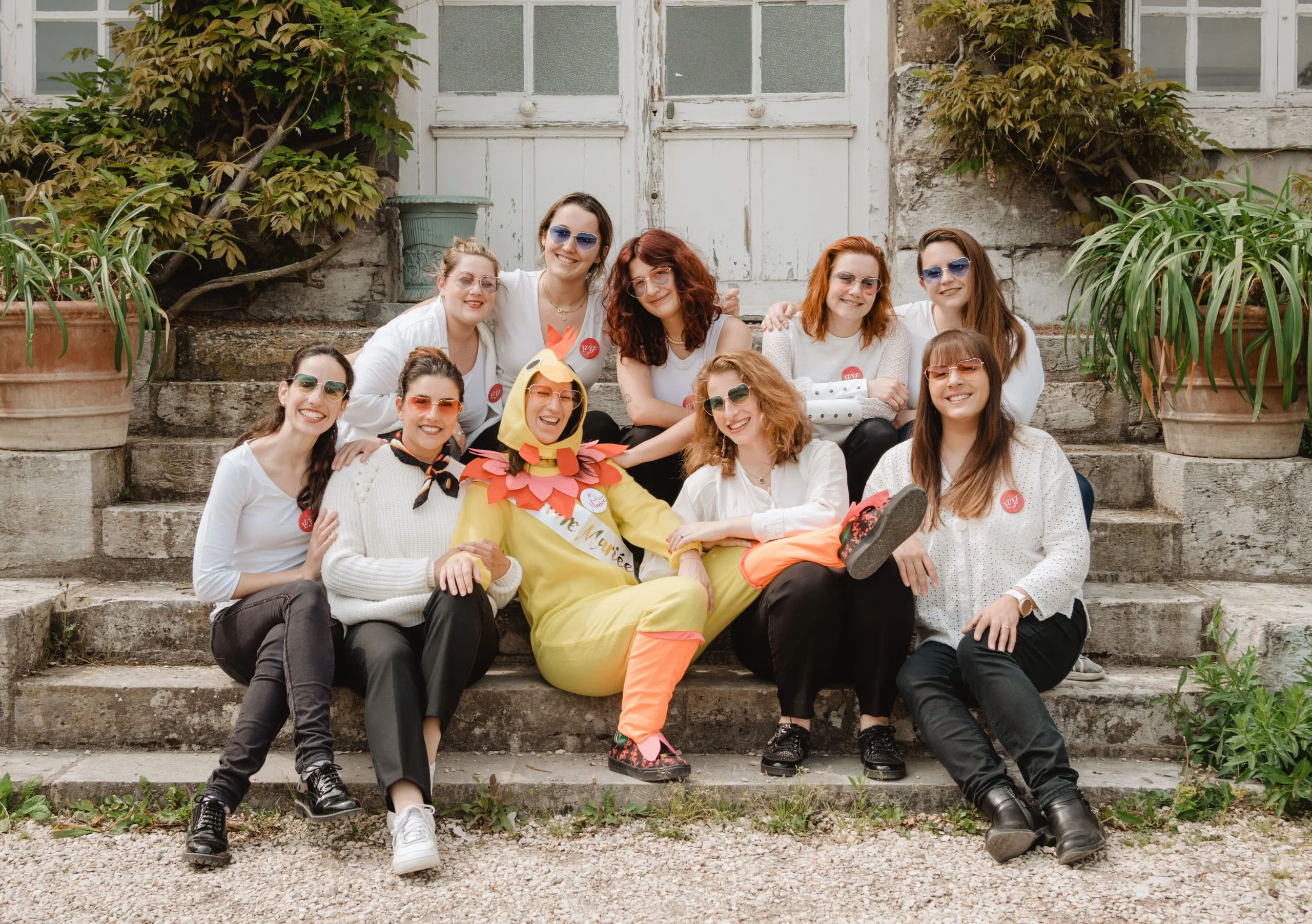 Groupe de femmes célébrant un EVJF à Rouen, posant sur des marches devant une maison ancienne, avec une participante déguisée en poulet pour un moment de fun et de complicité.