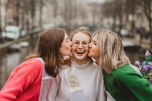 Groupe de femmes célébrant un EVJF à Amsterdam, avec des sourires éclatants et des bisous amicaux, en arrière-plan les canaux emblématiques de la ville.