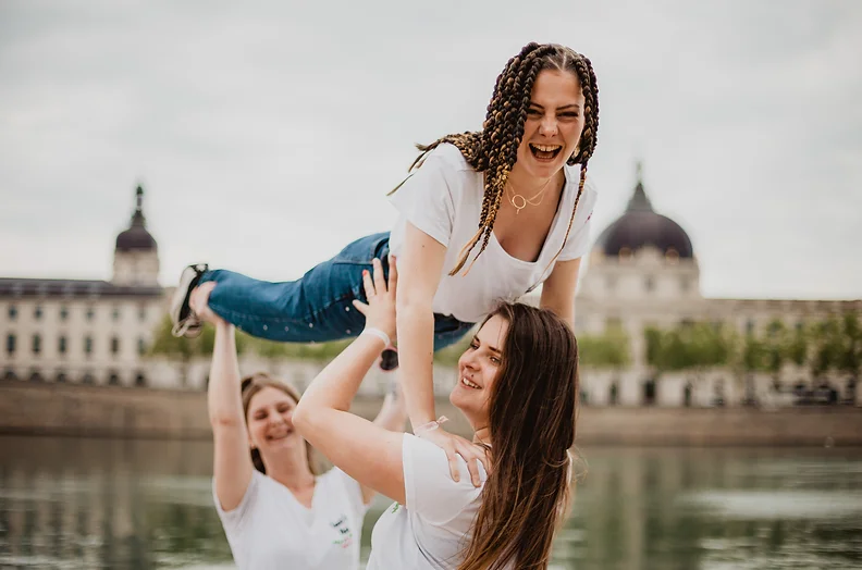 Groupe de femmes célébrant un EVJF à Lyon, avec une participante portée en riant devant le Rhône et l'Hôtel-Dieu en arrière-plan.