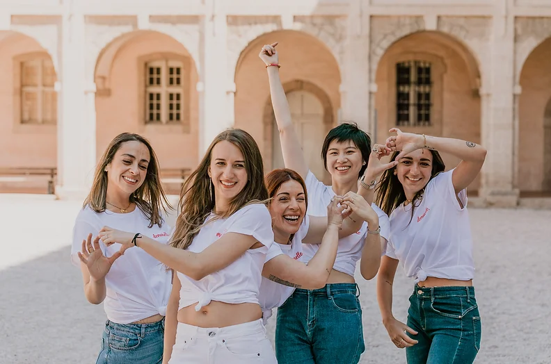 Groupe de femmes célébrant un EVJF à Marseille, réalisant un cœur avec leurs mains dans une cour ensoleillée entourée d'arches historiques.