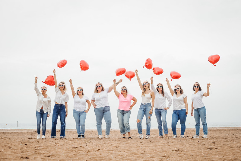 Groupe de filles EVJF sur la plage de La Baule brandissant des ballons en forme de cœur, lunettes et couronnes de fleurs