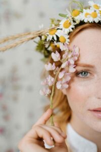 Femme portant une couronne de fleurs champêtres avec des marguerites et du blé, tenant une tige de fleurs roses devant son visage. Photo douce et poétique prise dans le cadre d’un atelier floral EVJF à Deauville.