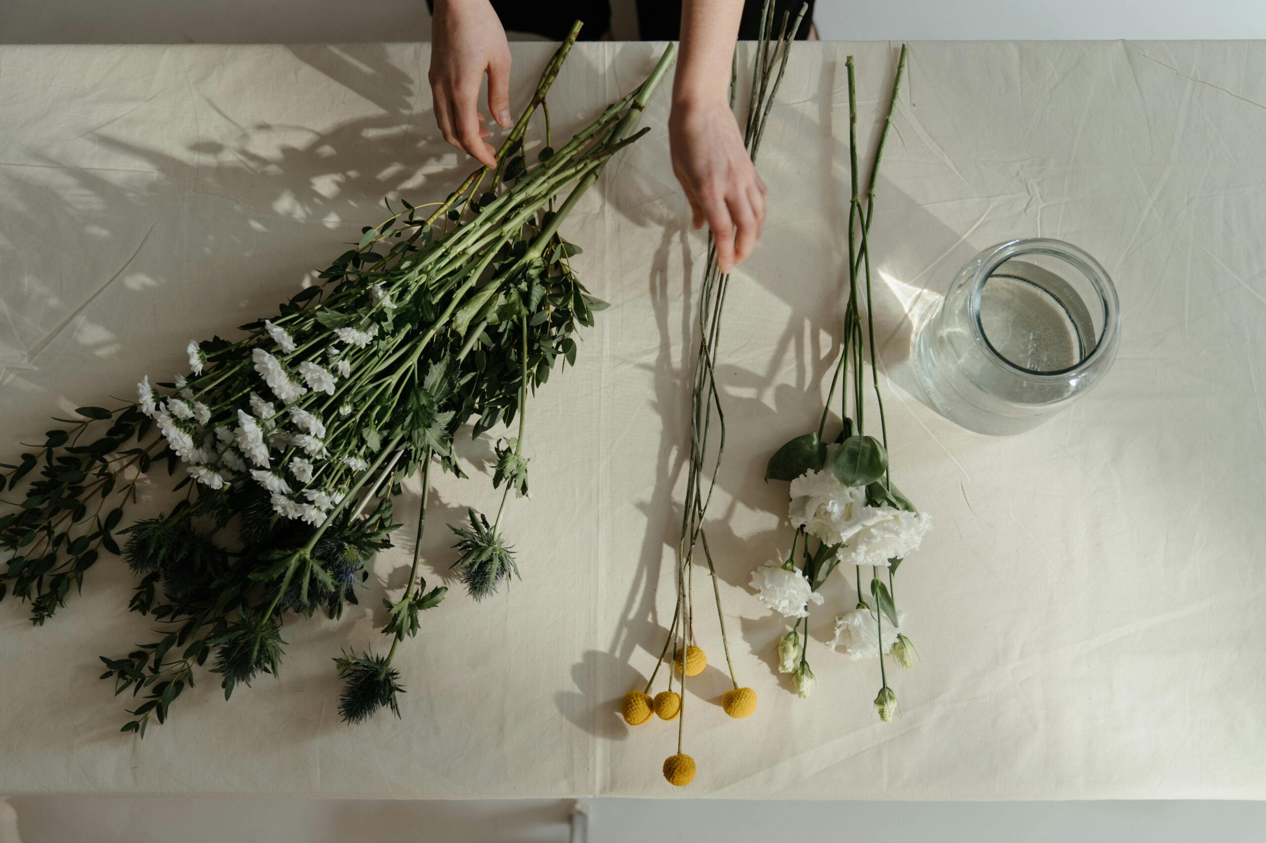 Préparation de compositions florales sur une table ensoleillée : des mains organisent différentes variétés de fleurs fraîches (craspedia, œillets, feuillage) pour un atelier floral EVJF à Deauville.