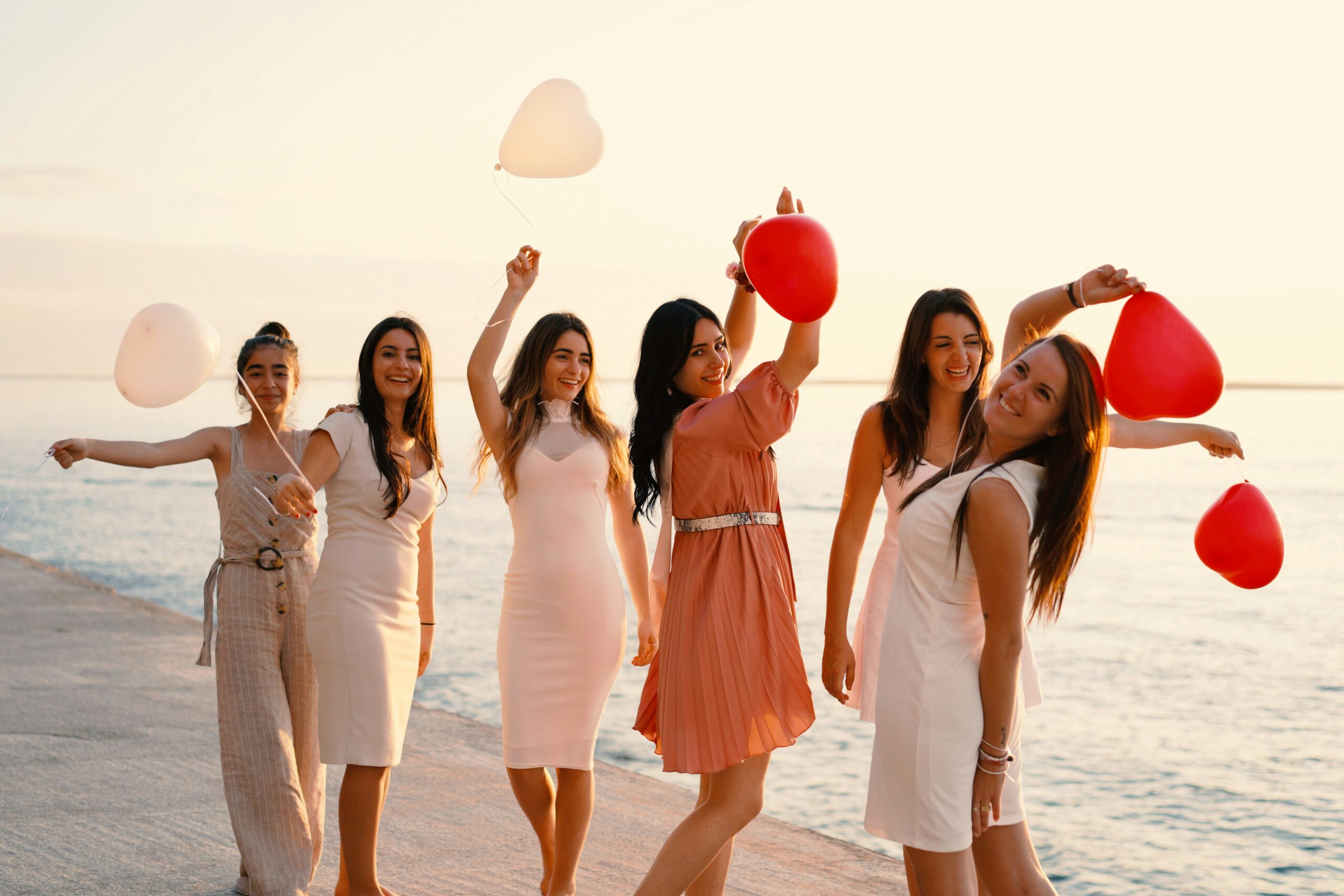 Groupe de femmes en tenue élégante avec des ballons en forme de cœur, célébrant un EVJF au bord de la mer à Marseille.