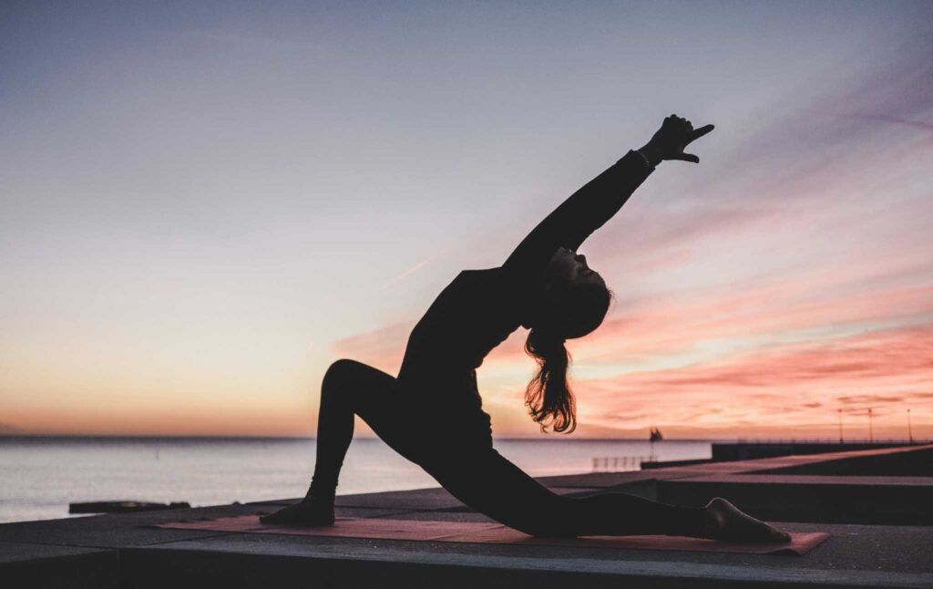 Une femme en posture de yoga au coucher du soleil, face à la mer, sur une plateforme en bord de plage.