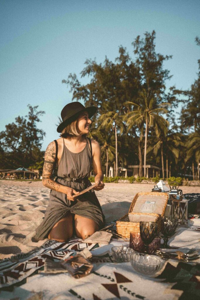 Panier pique-nique en osier ouvert sur une nappe au sol, entouré de verres, bocaux colorés et douce lumière naturelle, avec la mer en arrière-plan.