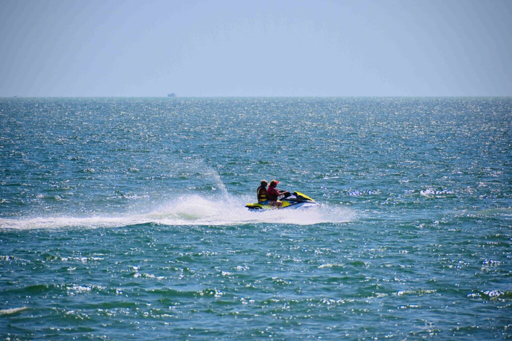 Groupe de jeunes femmes en gilets de sauvetage, souriantes à bord d’un zodiac en mer, pendant qu’une autre participante pilote un jet ski sur les vagues à proximité.