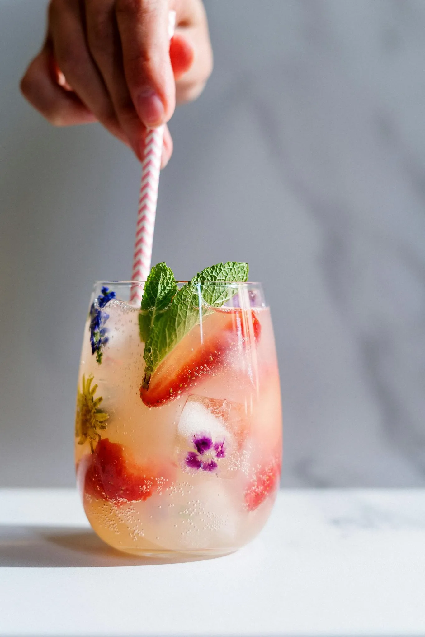 Main mélangeant un cocktail fruité avec des fraises, fleurs comestibles et feuilles de menthe dans un verre transparent – activité EVJF à Rouen