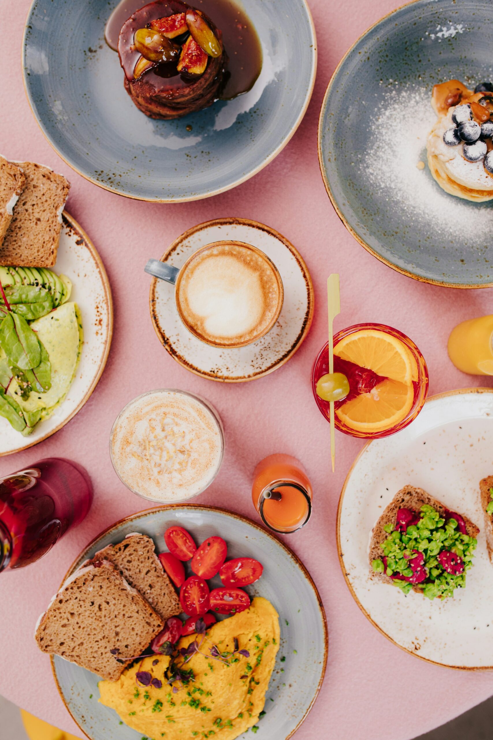 Table de brunch pour un EVJF à Biarritz avec omelette, avocado toast, pancakes, fruits frais, café et jus, sur fond rose élégant.