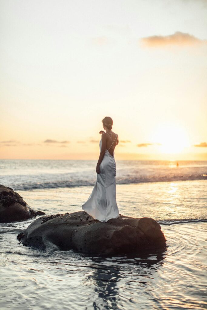 Jeune mariée de dos, debout sur un rocher entouré d’eau à La Baule, regardant vers l’horizon