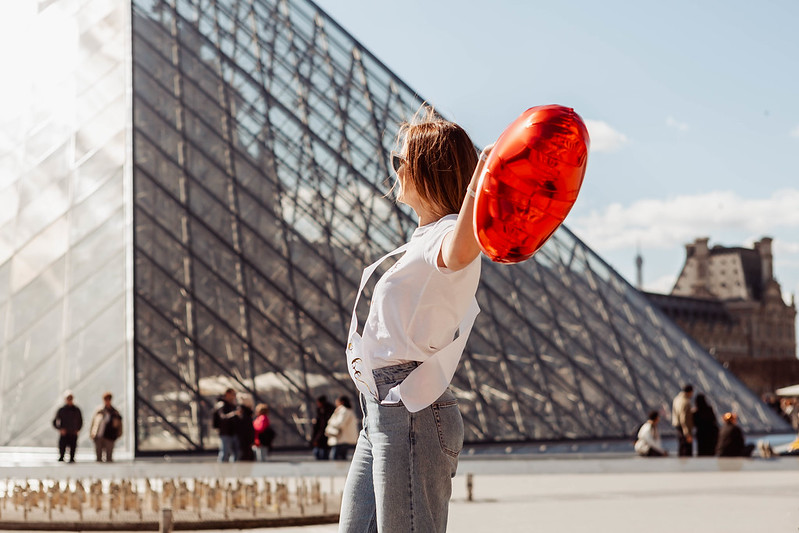 Future mariée de dos admirant la pyramide du Louvre à Paris, tenant un ballon coloré lors d’un shooting EVJF