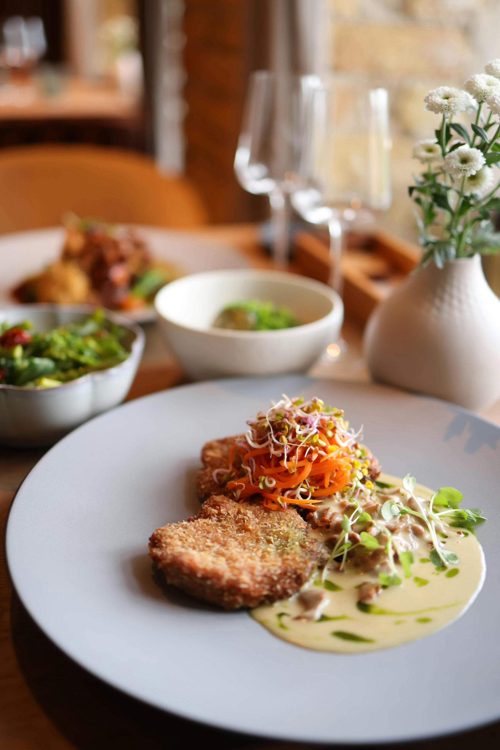 Plat raffiné dressé dans une assiette élégante, accompagné de légumes croquants et de sauce crémeuse, servi dans un restaurant à l’ambiance chaleureuse avec fleurs et verres à vin en arrière-plan.