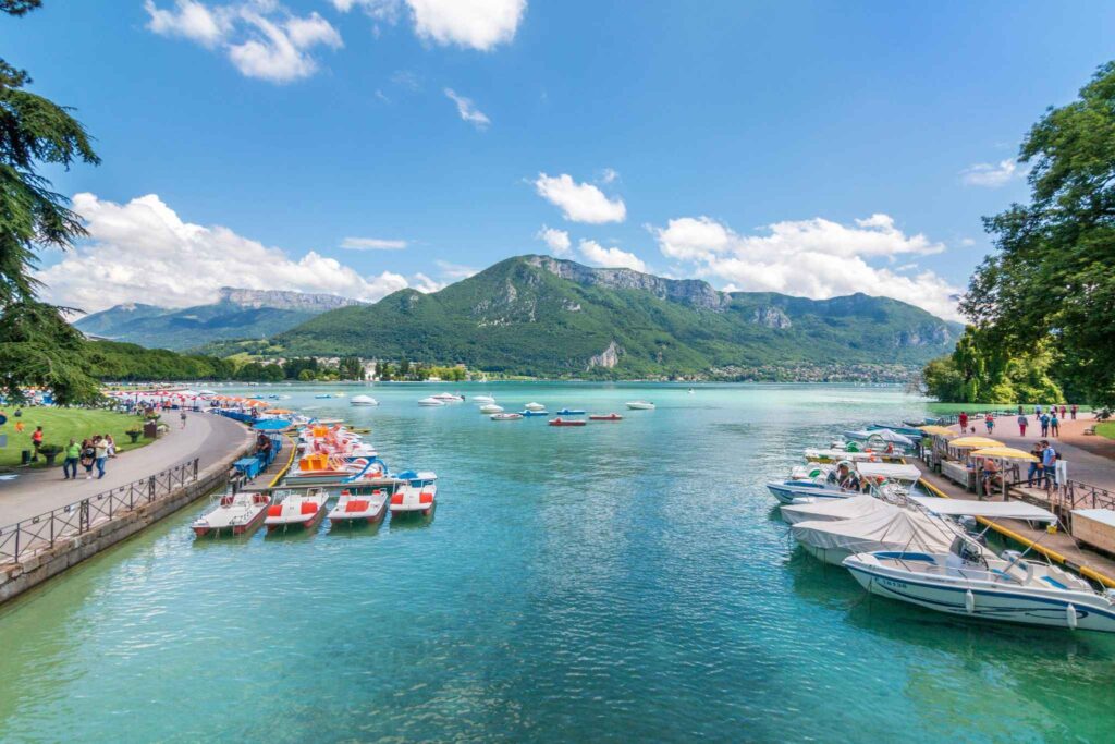 Vue panoramique du lac d’Annecy avec ses bateaux amarrés, ses eaux turquoise et les montagnes en toile de fond.