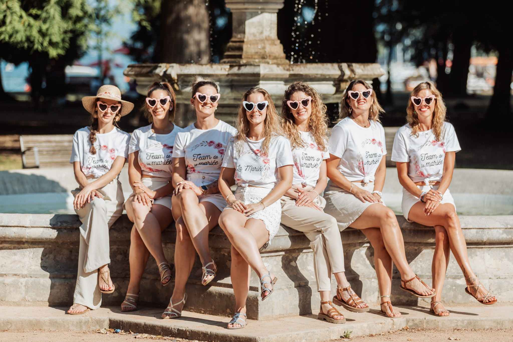 Groupe de femmes souriantes assises devant une fontaine dans un parc à Annecy, toutes vêtues de t-shirts EVJF "Team de la mariée", lunettes en cœur et tenues estivales.