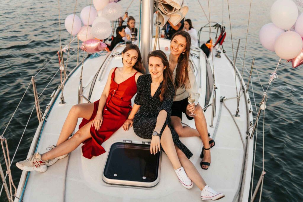 Jeunes femmes souriantes à bord d’un voilier décoré de ballons, profitant d’une croisière EVJF sur les eaux calmes au large de Deauville, par un beau temps de fin de journée.