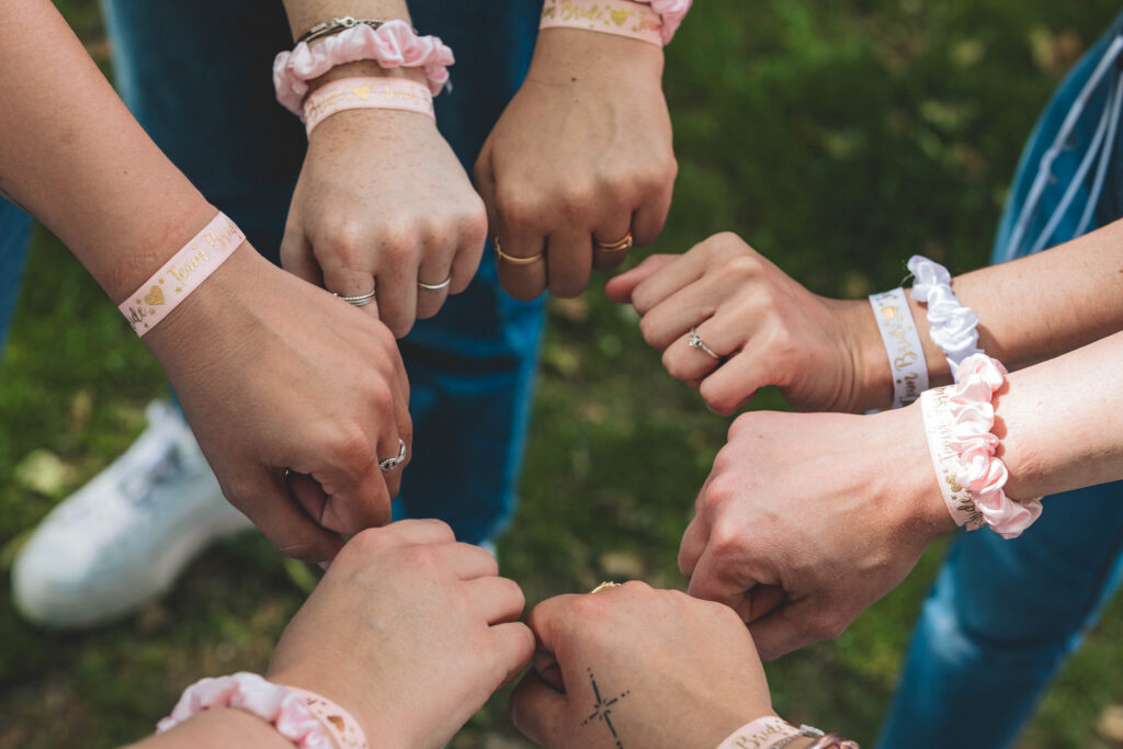 Cercle de mains de jeunes femmes portant des bracelets rose et blanc "Team Bride", serrant les poings en signe d’unité sur un fond d’herbe.