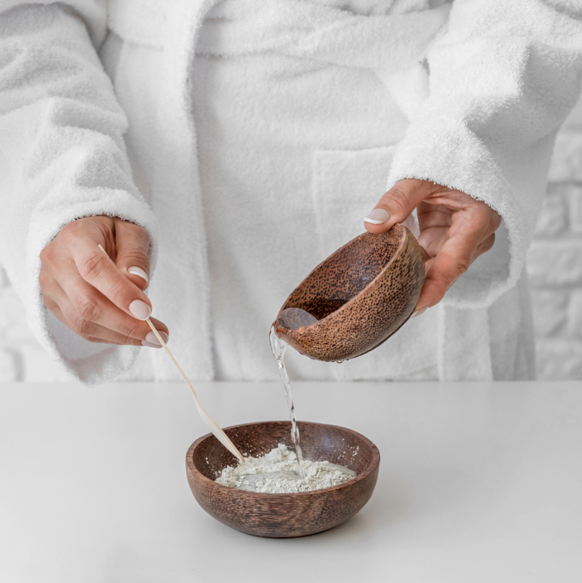 Femme versant de l’eau dans un récipient contenant de la poudre d’argile lors d’un atelier de création cosmétique