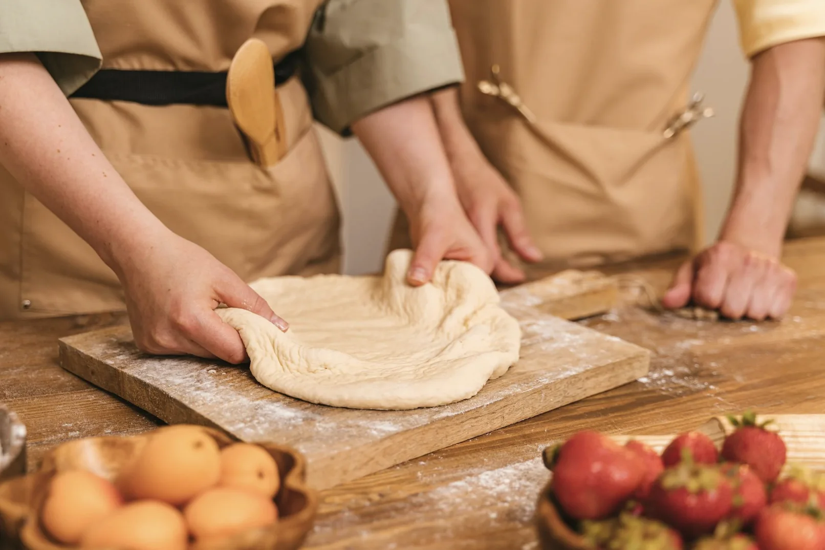 Deux personnes en tablier préparant une pâte à pizza sur un plan de travail en bois avec fruits frais à proximité