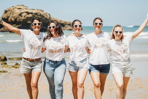 Groupe d’amies en t-shirts assortis et lunettes cœur marchant ensemble sur la plage lors d’un atelier DIY EVJF à Cabourg