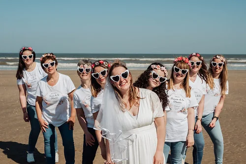 Future mariée entourée de ses amies en t-shirts blancs et lunettes cœur posant sur la plage lors d’un atelier DIY EVJF à Cabourg