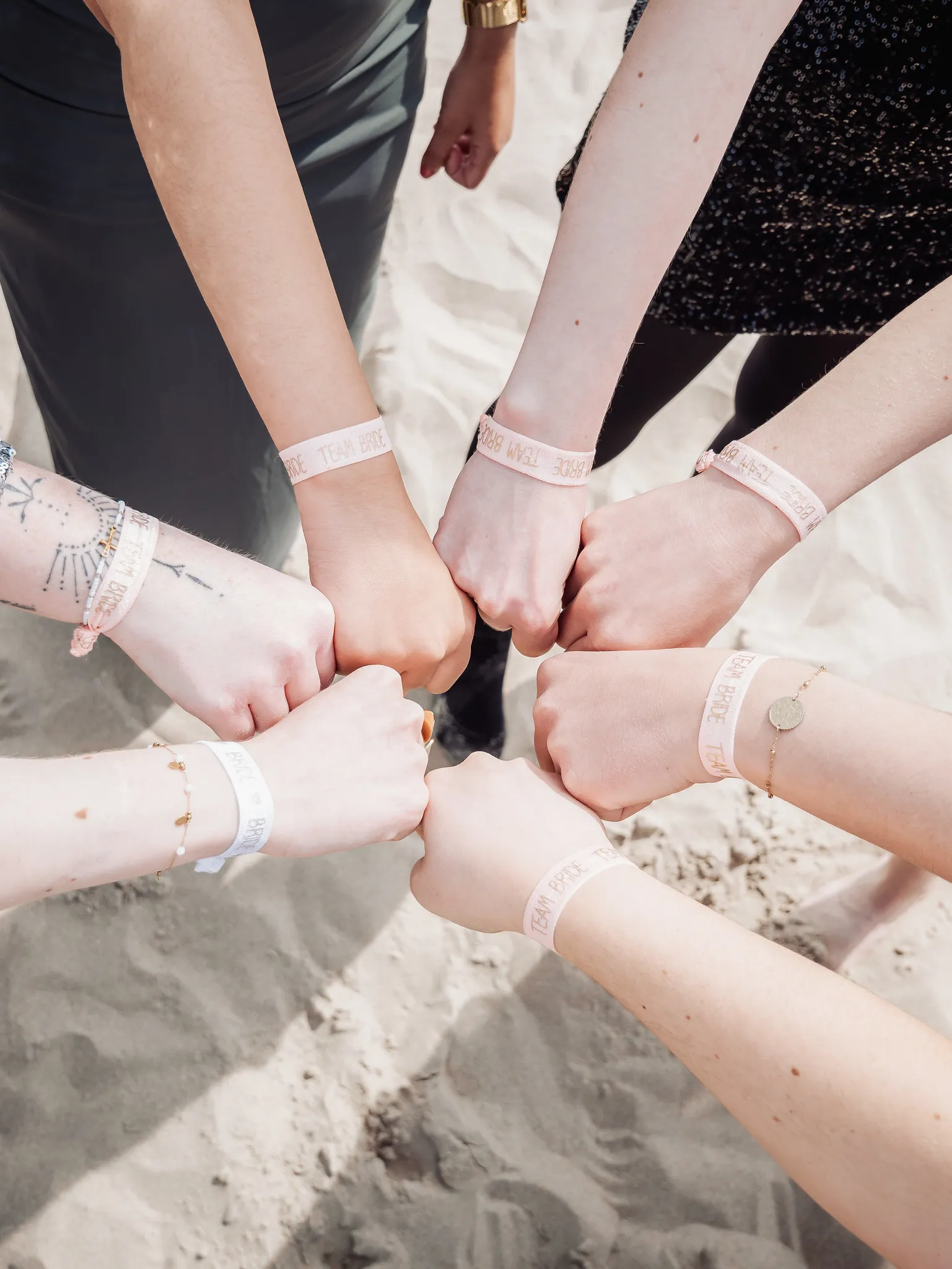 Huit mains de participantes à un EVJF formant un cercle sur la plage, portant des bracelets "Team Bride"