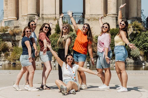 Groupe de jeunes femmes souriantes, bras levés, posant devant un monument lors d’une chasse au trésor EVJF à Montpellier.