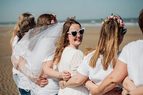 future mariee souriante avec voile et lunettes coeur entouree de ses amies sur la plage de cabourg pendant un evjf