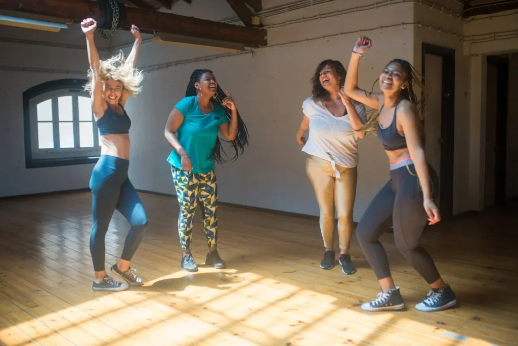 Femmes en tenue de sport dansant et riant ensemble dans un studio lumineux à Paris lors d’un EVJF.Image de femmes riant et dansant ensemble dans un studio lumineux à Paris lors d’un cours de danse EVJF.