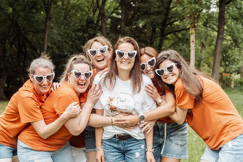 groupe de femmes en t shirt orange entourant la future mariee avec des lunettes coeur blanches