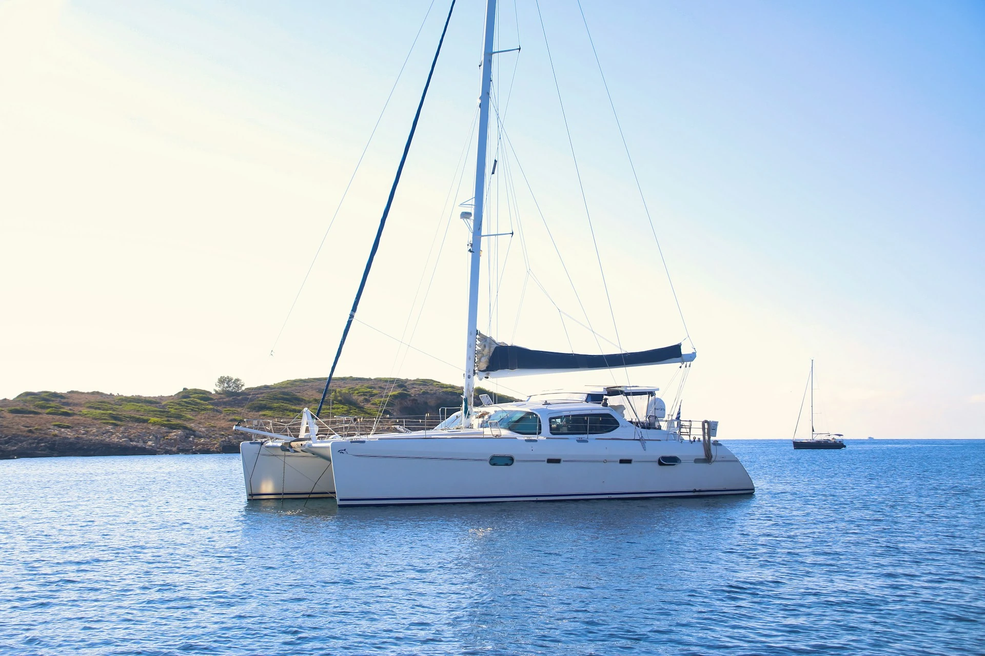 Grand catamaran blanc au mouillage près d’une côte rocheuse et verdoyante au soleil.