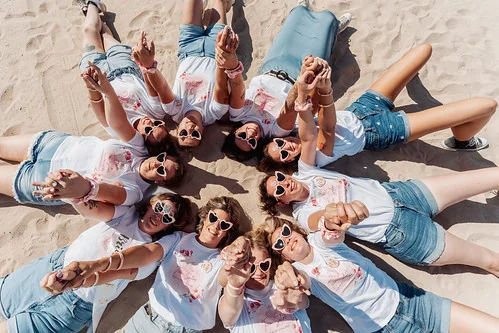 groupe de femmes en short et t shirts blancs allongees en cercle sur le sable levant les bras lors dun evjf a la rochelle