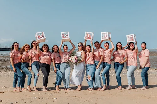 groupe de femmes en t shirts roses et jeans celebrant un evjf sur la plage avec la mariee tenant un bouquet