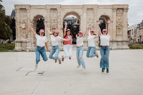 Groupe de femmes en jeans et t-shirts blancs sautant ensemble devant un arc historique lors d’un clip video evjf à Reims