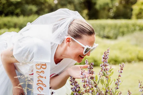 Femme avec voile et écharpe bride team sentant des fleurs dans un jardin lors d’un clip video evjf à Reims