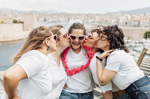 evjf-marseille-bisous-mariee future mariee souriante avec couronne et lunettes coeur embrassee par ses amies