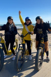 Trois amies souriantes sur des trottinettes electriques jaunes au bord de la plage lors d un EVJF a La Baule.