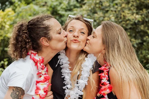 femme embrassee sur les joues par deux amies portant des colliers de fleurs