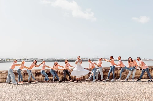 groupe de femmes en t shirts roses et jeans tire la mariee en robe blanche qui sourit sur un muret face a la mer a la rochelle