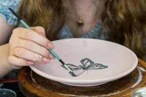 Main féminine peignant un papillon noir sur une assiette en céramique rose.