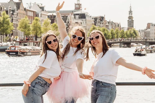 trois amies souriantes posent devant un canal d’amsterdam pendant un jeu de piste evjf paranormal dans une ambiance joyeuse