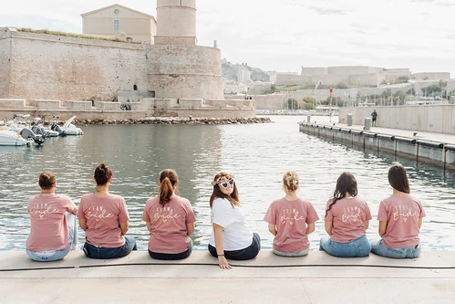 Groupe d’amies assises au bord du Vieux-Port à Marseille pendant un jeu de piste EVJF paranormal, vue sur le fort Saint-Jean