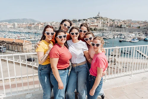 Groupe d’amies souriantes en t-shirts colorés posant au Vieux-Port de Marseille pendant un jeu de piste EVJF paranormal