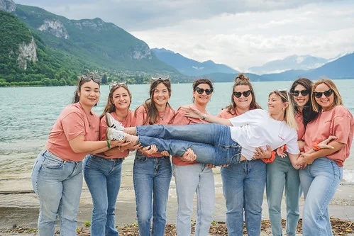 Groupe de filles souriantes portant une amie dans les bras au bord du lac, ambiance koh lanta evjf annecy