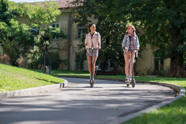 Deux amies roulent en trottinette électrique dans un parc partageant une balade conviviale.