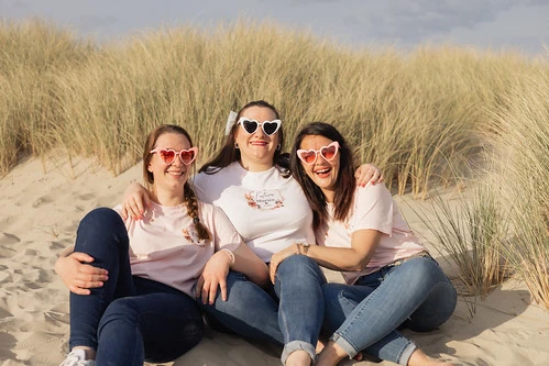 nappe blanche pique nique appareil photo bouquet de fleurs et bouteille de vin trois amies assises dans les dunes portant des lunettes coeur lors d’un evjf au touquet paris plage