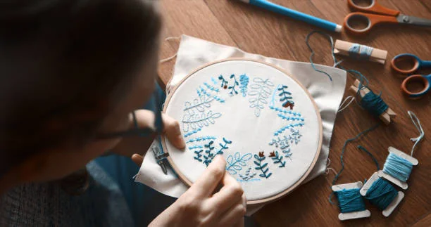 Femme réalisant une création textile lors d’un atelier broderie , avec fils bleus et motif floral sur tambour à broder.