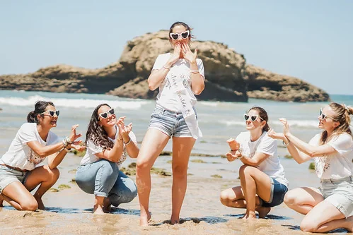 Groupe d’amies sur la plage entourant la future mariée pendant un clip vidéo EVJF à Biarritz sous un ciel ensoleillé.