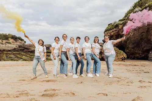 Groupe de femmes souriantes sur la plage de Biarritz tenant des fumigènes colorés pendant un clip vidéo EVJF.