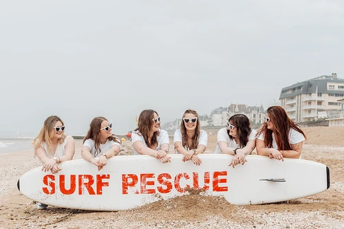 six amies en t shirts blancs posant sur la plage de cabourg derrière une planche de surf pour des defis et gages evjf