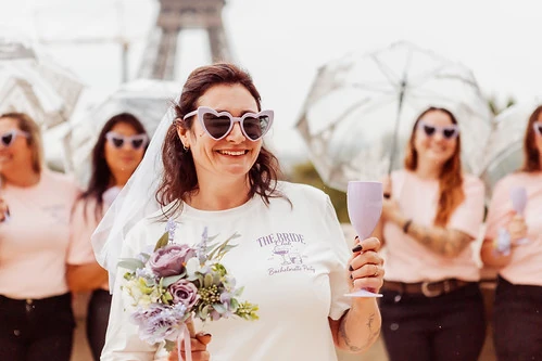 Future mariée souriante tenant un bouquet et un verre, entourée de ses amies pendant un escape game EVJF à Paris près de la tour Eiffel